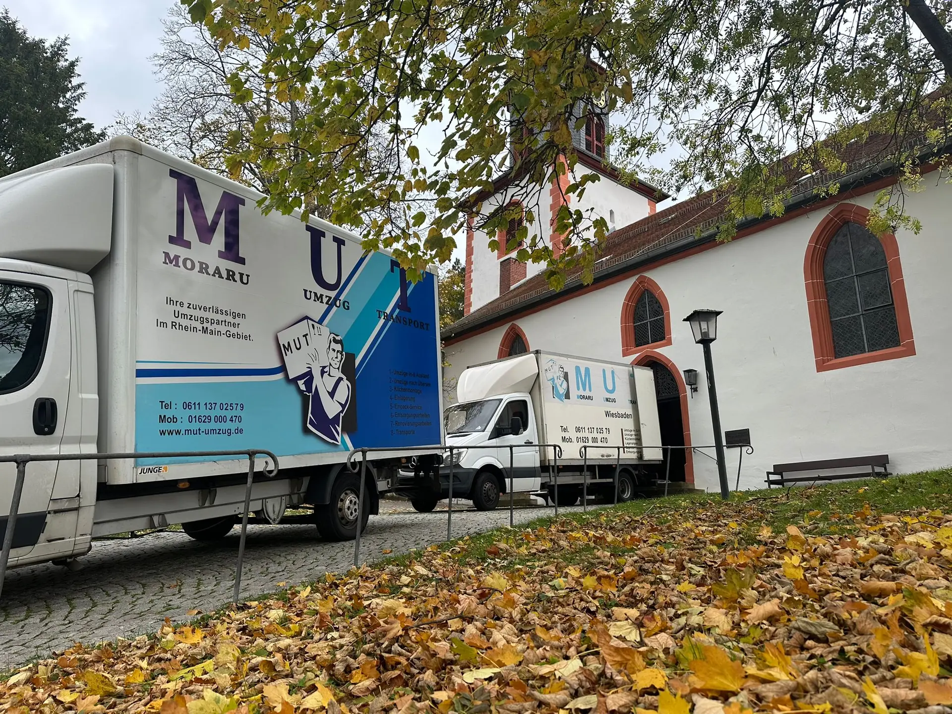 MUT-Umzugswagen vor einer Kapelle im Herbstlaub