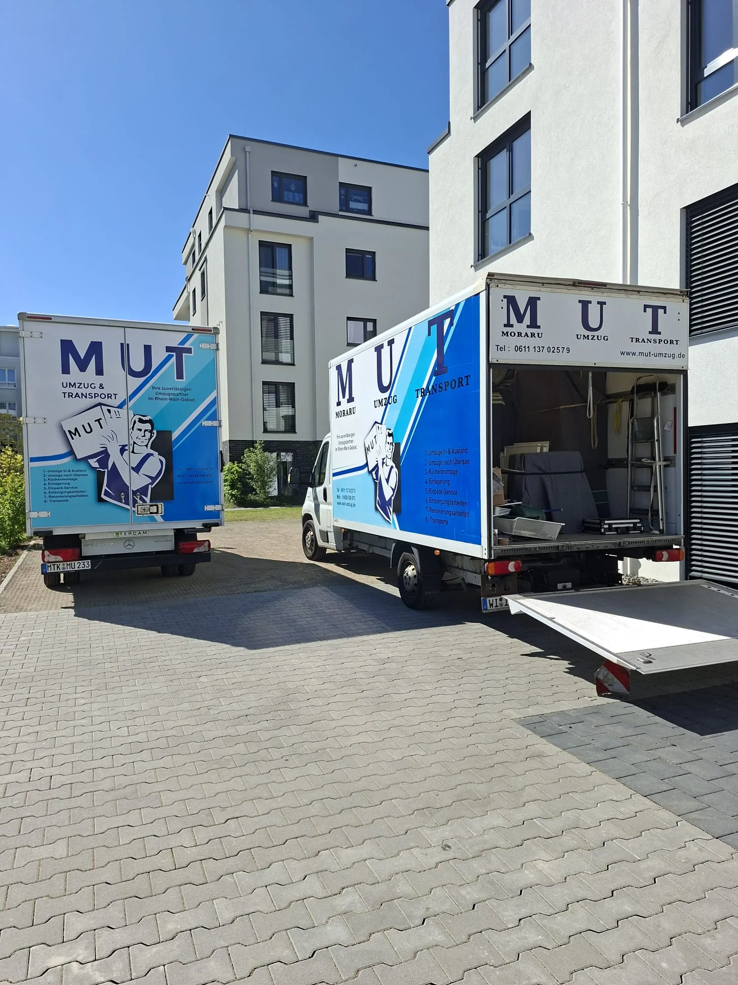 Zwei MUT-Umzugswagen mit geöffneter Ladebordwand vor einem Wohnhaus