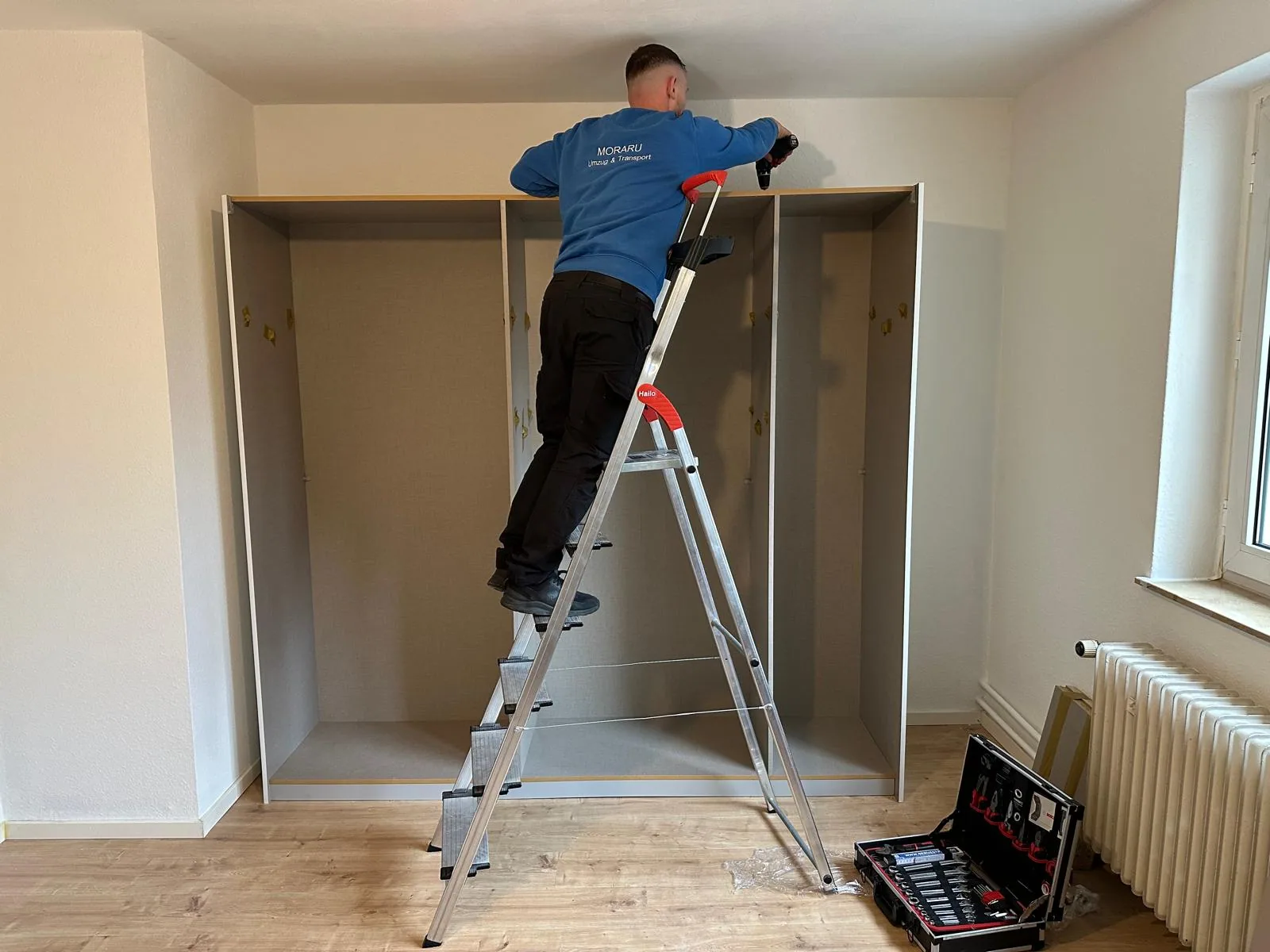 MUT-Mitarbeiter baut auf einer Leiter einen Kleiderschrank ab, daneben der geöffnete Werkzeugkoffer